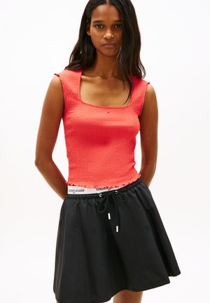 Femme portant un haut corail ajusté sans manches et une jupe noire à cordon avec une ceinture blanche visible portant la marque Tommy Jeans.