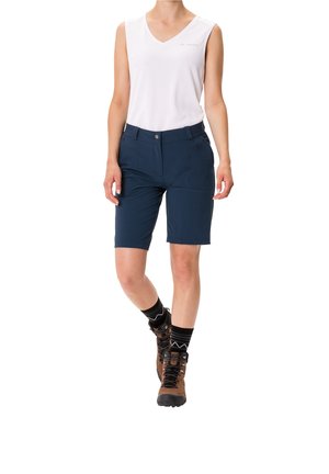 Marineblauwe wandelshorts met voorzakken, gecombineerd met een witte mouwloze top. Het model draagt bruine wandelboots en gestreepte zwarte sokken.