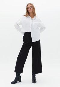 Camicia bianca a maniche lunghe con taschino, abbinata a pantaloni larghi neri e stivali alla caviglia neri, che evidenziano una texture liscia e una vestibilità su misura.