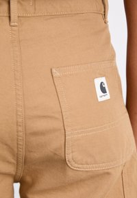 Beige cotton work pants feature a large side pocket and a stitched logo label. The fabric has a smooth texture with durable stitching details.