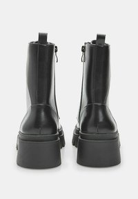 Un paio di stivaletti neri in pelle con tacchi a piattaforma chunky, cerniera posteriore e linguette, mostrati da dietro su uno sfondo bianco.