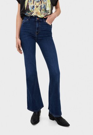 Persoon die een donkerblauwe flarebroek met hoge taille draagt, zwarte enkellaarsjes met puntige neus en een zwart T-shirt met een Egyptisch thema grafische print.