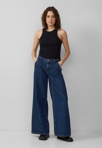 Haut sans manches noir avec de petites décorations, associé à un jean bleu taille haute et large. Le mannequin a les mains dans les poches et porte des chaussures noires.