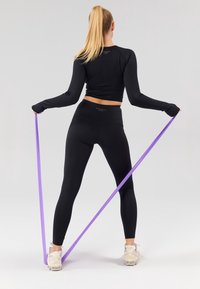 Czarna bluzka z długim rękawem i legginsy typu high-waisted, trzymająca fioletową taśmę oporową. Jasne buty sportowe. Prosty, funkcjonalny design.