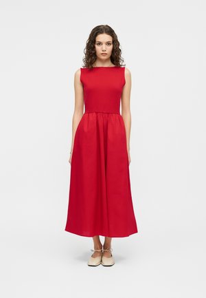 Mujer con cabello rizado, vestida con un vestido midi rojo sin mangas y zapatos planos beige con lazo en el tobillo, de pie contra un fondo blanco liso.