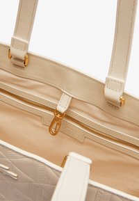 Sac à tote beige avec surface texturée, fermeture éclair dorée et poches intérieures. Doté de poignées solides et d'une doublure lisse.