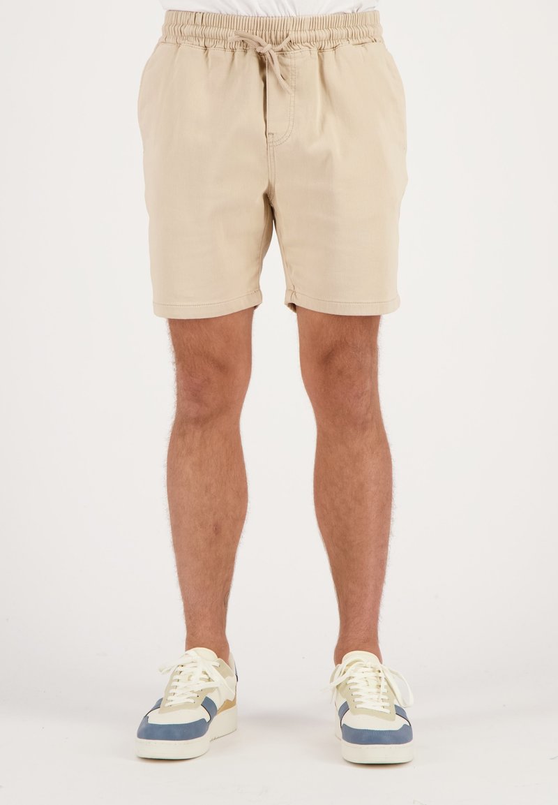 Beige shorts met een elastische tailleband en een trekkoord. Stof lijkt glad. Draag met lichtgekleurde sneakers met blauwe en beige accenten.