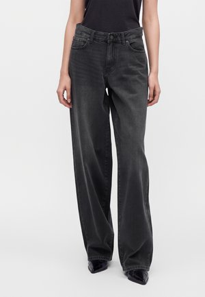 Vrouw die losse zwarte hoge taille jeans met wijde pijpen en zwarte puntige schoenen draagt, staand tegen een effen lichte achtergrond.
