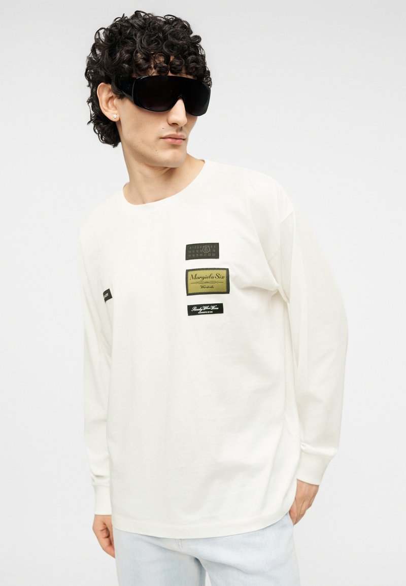 T-shirt blanc à manches longues avec divers patchs en tissu noir et or. Texture lisse, coupe décontractée, porté avec des lunettes de soleil noires oversize.
