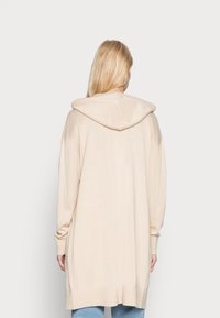 Beżowy kardigan z kapturem, długimi rękawami i ściągaczami przy mankietach, o luźnym kroju i bocznych rozcięciach zapewniających swobodę ruchu.