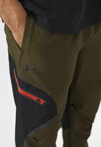 Pantalons de sport vert olive avec des accents noirs, dotés d'une fermeture éclair orange sur une poche avant et d'un logo sur la cuisse. Tissu lisse et extensible.