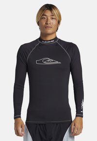 Černý dlouhý rukáv rashguard s plochými švy, vysokým límcem a bílým logem na hrudi. Vyrobeno z pružné, vlhkost-odvádějící látky.