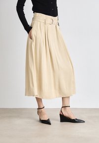 Beige, redőzött midi szoknya széles derékpánttal és ezüst csattal, fekete, hegyes orrú wedge cipővel párosítva. Simább anyag textúra.