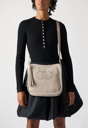 Borsa a tracolla in pelle beige con logo perforato, dettaglio con frange e tracolla regolabile, abbinata a un vestito nero a maniche lunghe a coste.