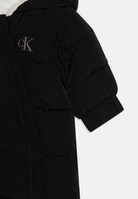 Veste matelassée noire avec capuche, présentant une texture matelassée, une fermeture éclair et un petit logo brodé sur la poitrine.