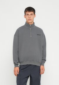 Sudadera gris con cremallera en el cuello y un corte relajado, hecha de una tela suave. Cuenta con puños y bajo acanalados, con un sutil logo en el pecho.