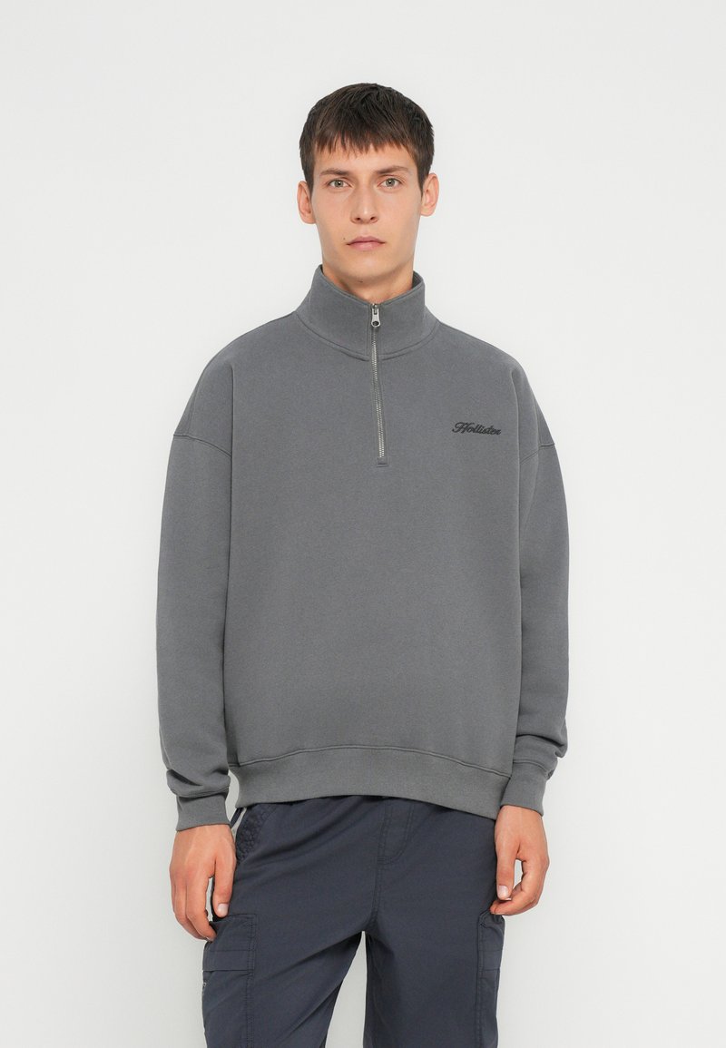 Sudadera gris con cremallera en el cuello y un corte relajado, hecha de una tela suave. Cuenta con puños y bajo acanalados, con un sutil logo en el pecho.