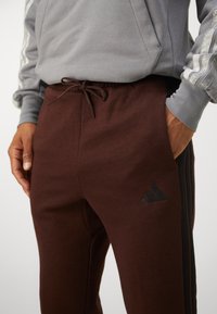 Barna melegítőnadrág zsinóros derékkal, puha textúrával, fekete oldalcsíkokkal, és fekete Adidas logóval a bal combon. Szürke kapucnis pulóver látható felette.