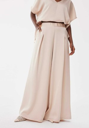 Femme portant un pantalon beige à jambes larges et un haut ample assorti, main dans la poche, debout sur un fond clair.