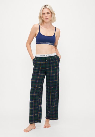 Blonda sieviete stāvot basām kājām, ģērbusies tumši zili sporta krūšturī un zaļā kvadrātveida pidžamu biksēs ar Polo Ralph Lauren zīmolu jostu.