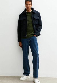 Mörkblå fleecejacka med två bröstfickor, olivgrön ribbad tröja, blå jeans och vita sneakers mot en neutral bakgrund.