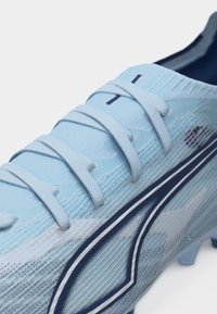 Light blue cleat with textured upper, white laces, dark blue logo on side, and studded sole, shown in close-up detail.