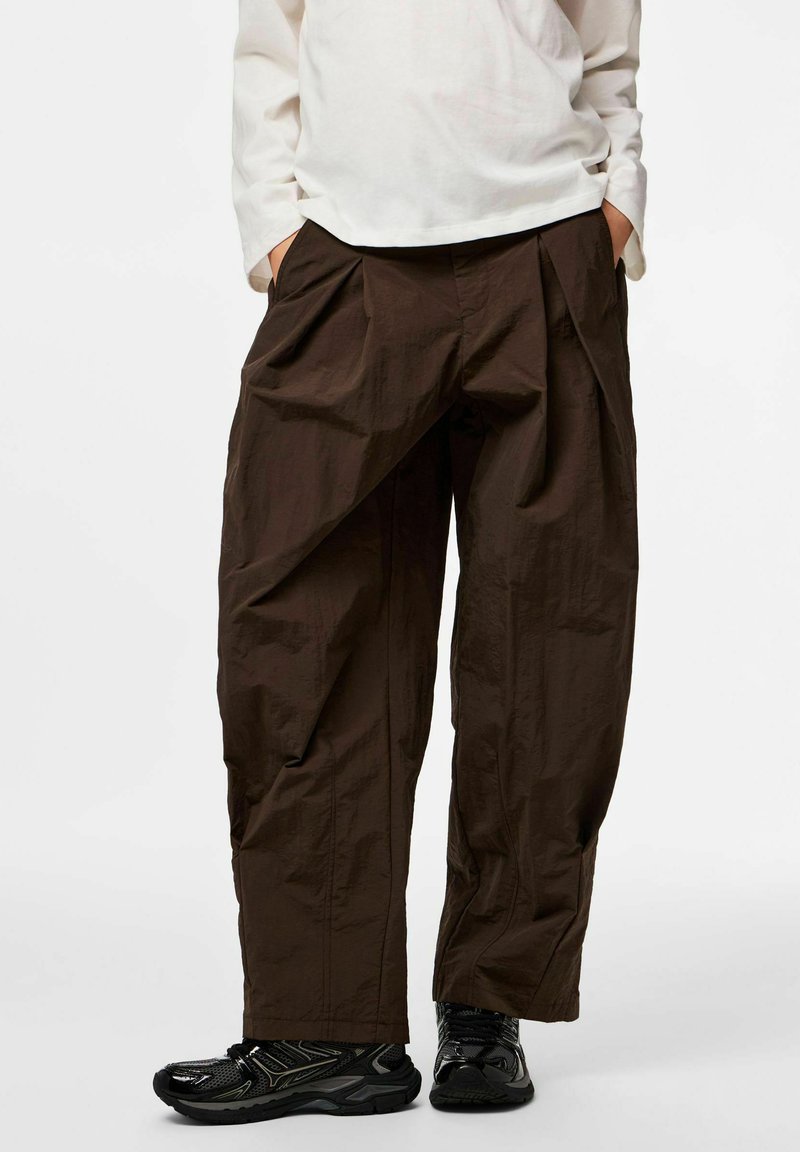 Osoba nosící volné hnědé kalhoty, černé tenisky a bílé tričko s dlouhým rukávem, s rukama v kapse, stojící na jednobarevném pozadí.