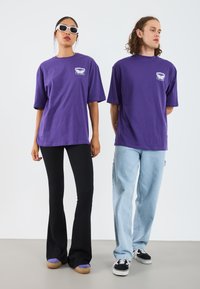 Camisetas de algodón moradas de manga corta con un gráfico en el pecho, combinadas con pantalones de campana negros y jeans de color azul claro. El calzado incluye zapatillas de plataforma y zapatillas de skate.