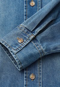 Blaues Jeanshemd-Manschettenende mit abgerundeter Kante, verstärkten Nähten und zwei hellbraunen Knöpfen mit jeweils vier Löchern. Weiche Textur mit verblassten Akzenten.