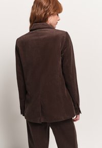 Blazer in corduroy marrone con texture a coste verticali, colletto a rever e spacco posteriore. Presenta una vestibilità tailored e polsini con bottoni.