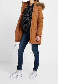 Brown parka with a faux fur hood, zip closure, and drawstring details. Worn over a black polka dot shirt and paired with blue jeans and white sneakers.
