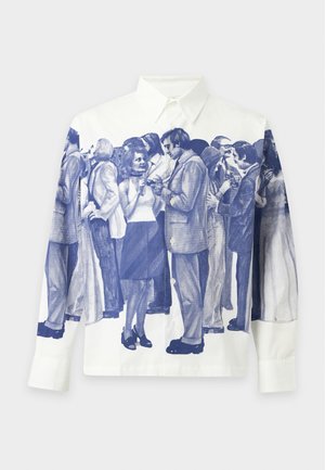 Mand iført hvid skjorte med blå folkemængdeprint, lyseblå jeans, sorte sko og et sølvarmbånd, stående mod en ensfarvet baggrund.