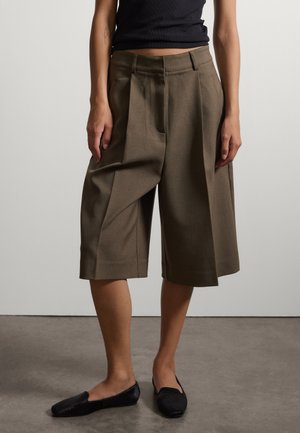 Person trägt lockere braune knielange Shorts, schwarzes ärmelloses Top und schwarze flache Schuhe, steht auf grauem Boden vor weißem Hintergrund.