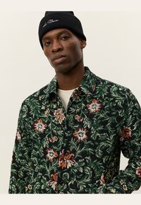 Floral-patterned shirt in dark green with red, orange, and white flowers. The fabric has a textured finish, featuring a collar and buttons.