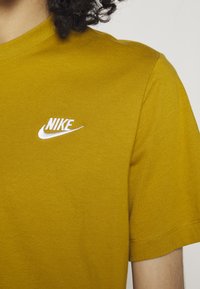 Majica iz bombaža v gorčično rumeni barvi ima bele vezenine Nike logotipa na levem prsnem delu. Mehka tekstura in krojen vratni izrez.