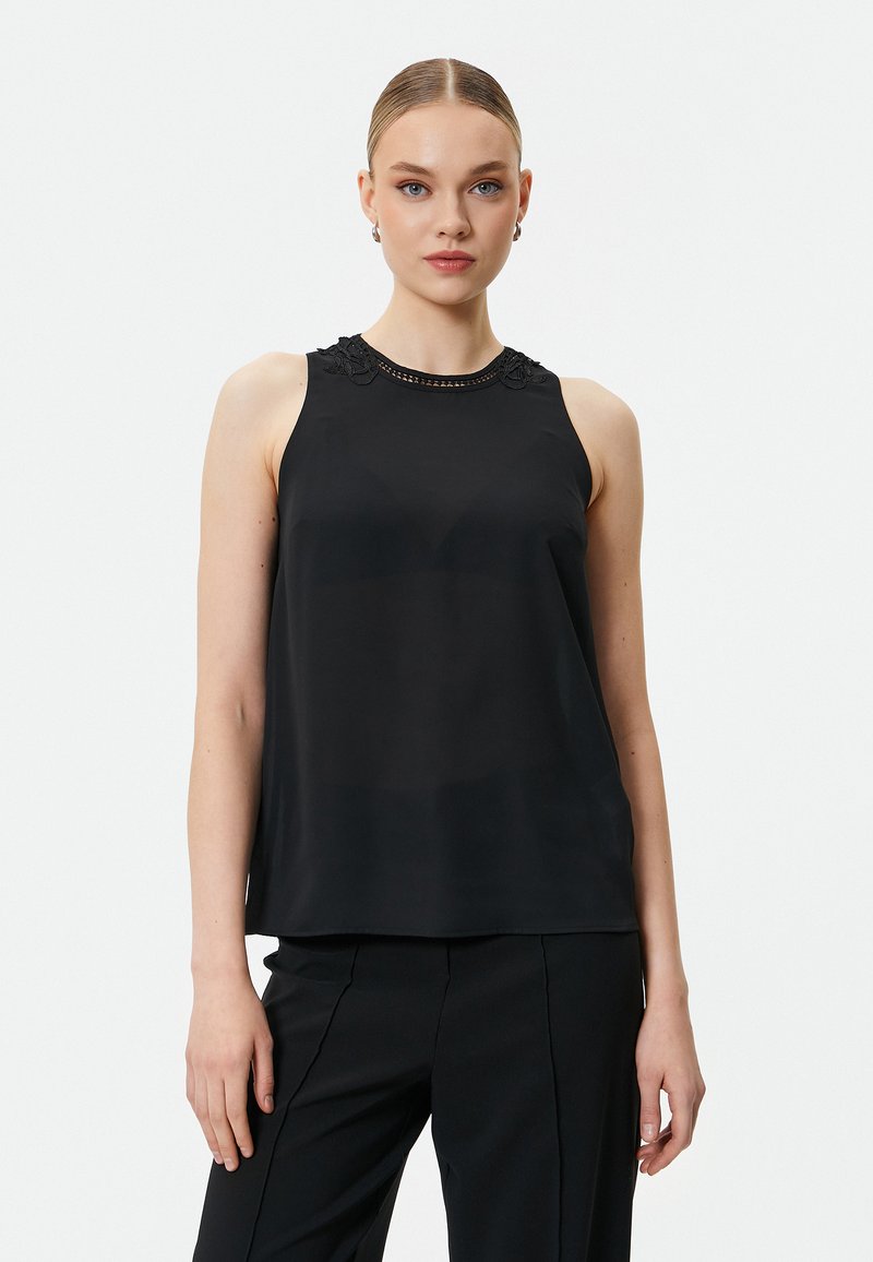 Top negro sin mangas con un cuello alto, que presenta un intrincado detalle de encaje y una tela suave y ligera. Combinado con pantalones negros a medida.