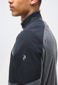 Άνδρας με μαύρο και γκρι υφασμάτινο μπουφάν με ψηλό γιακά, εμφανιζόμενος από το πλάι με έμφαση στους ώμους και την περιοχή του λαιμού.