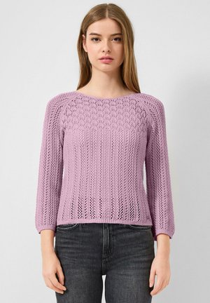 Jonge vrouw die een lila gebreide trui draagt met een gestructureerd patroon en driekwart mouwen, gecombineerd met zwarte jeans tegen een effen achtergrond.