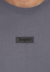 Grå T-shirt med rund halsudskæring, der har en sort læderpatch med "Dropsize" i præget tekst. Glat, ensartet stof tekstur.