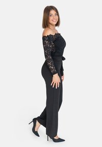 Top off-shoulder in pizzo nero, pantaloni neri aderenti e décolleté neri a punta. Tessuto liscio con un dettaglio legato in vita.