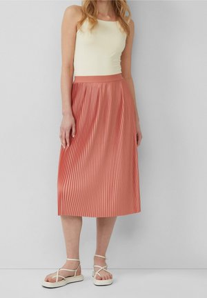 Kobieta ubrana w kremowy top bez rękawów, koralową plisowaną spódnicę midi i białe sandały na platformie, stojąca na tle szarej ściany.