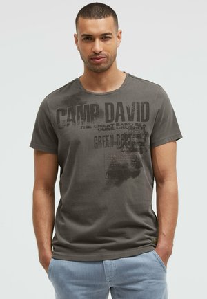 Man wearing a dark gray Camp David graphic t-shirt and light blue pants, standing with hands in pockets against plain background.