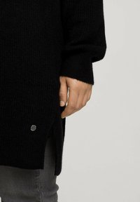 Hand auf der Hüfte ruhend, trägt einen schwarzen Strickpullover mit seitlichem Schlitz und kleinem runden Metalllogo über grauen Hosen.