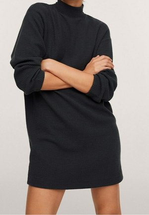 Personne portant une robe pull noire texturée à manches longues, les bras croisés, debout devant un fond clair uni.