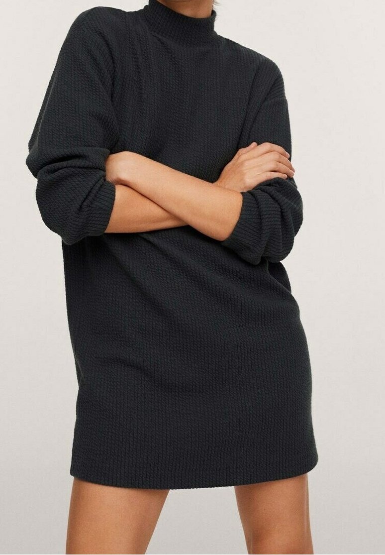 Personne portant une robe pull noire texturée à manches longues, les bras croisés, debout devant un fond clair uni.