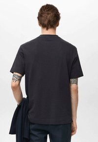 Tricou în navy închis, cu mâneci scurte, guler rotund și un material texturat. Tatuaj vizibil pe braț, însoțit de un pulover pliat.