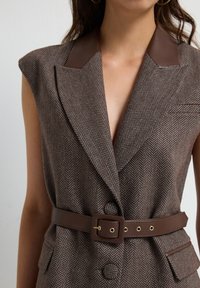 Brun houndstooth vest med læderkrave, bælte i taljen og to forlommer, med dyb V-udskæring og strukturerede skuldre.