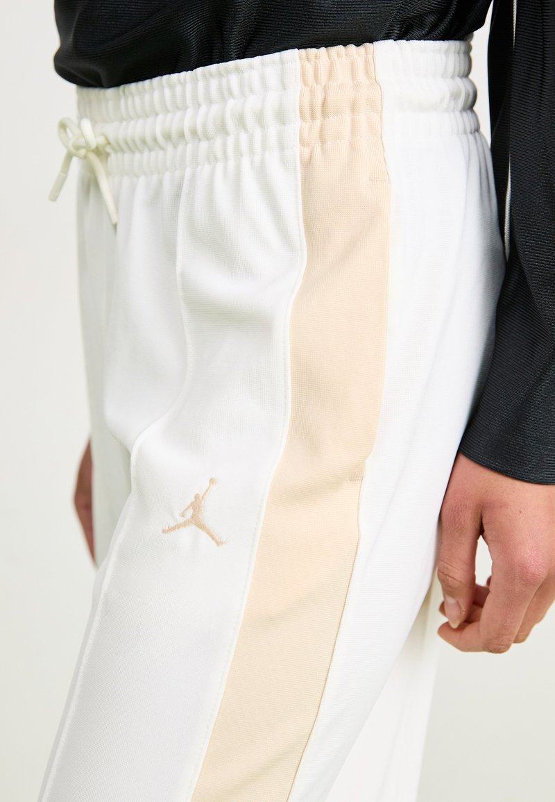Pantalones deportivos blancos con una franja lateral beis, cintura elástica con cordón y un pequeño logo bordado en la pierna izquierda.