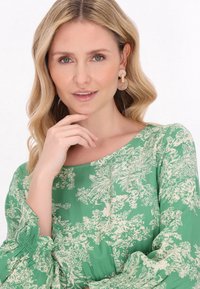 Zöld virágos ruha hosszú, puffos ujjal és széles rugalmas mandzsettával. Krém színű, világos virágmintával rendelkezik. A modellen átlátszó, kerek fülbevalók vannak.