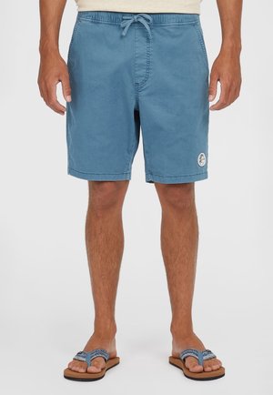 Mann trägt blaue Shorts mit Kordelzug und blaue gemusterte Flip-Flops, steht vor einem schlichten weißen Hintergrund.
